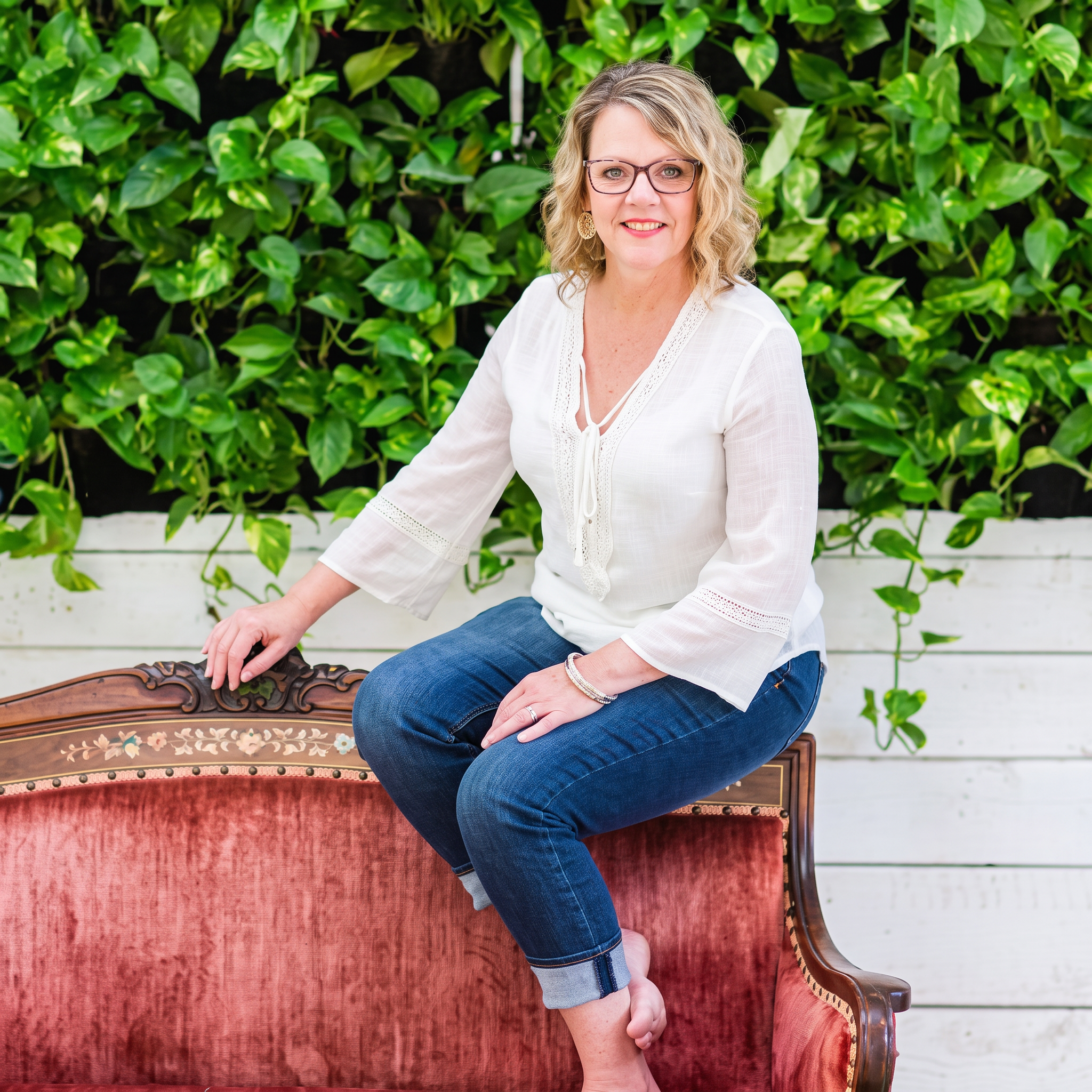 Jaynee McGinn, designer and owner of Blue Heron Home + Gardens, seated on an antique rose velvet settee against a wall of ivy